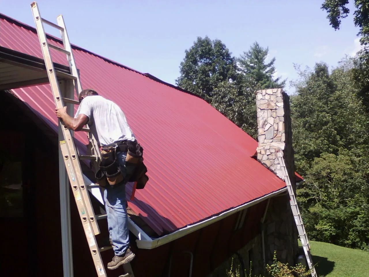 Roofer on a ladder beside a red metal roof and stone chimney — Soffit & Fascia Repair by Roofing Brothers in Table Rock